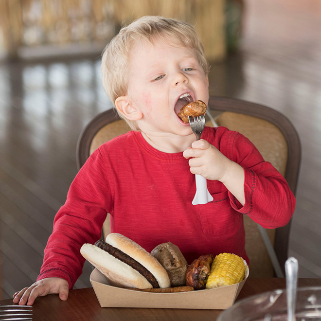 A small boy eating a sausage with a plate full of BBQ food